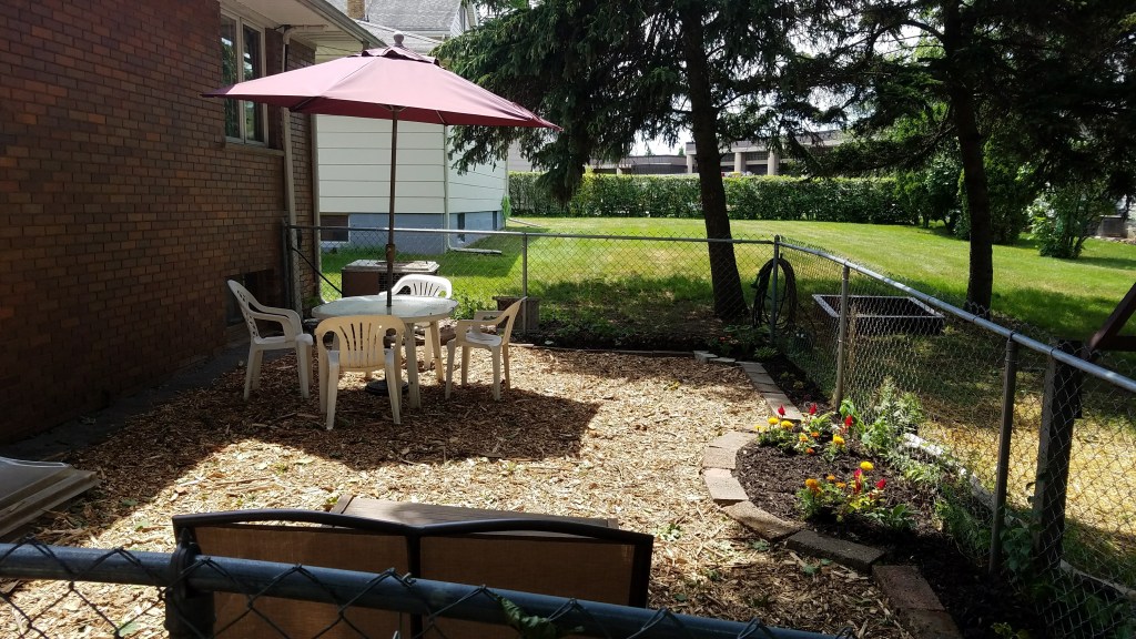 Patio inside a chain-link fence with mulch groundcover, white table and chairs under a red umbrella, and flowerbeds along the edges.