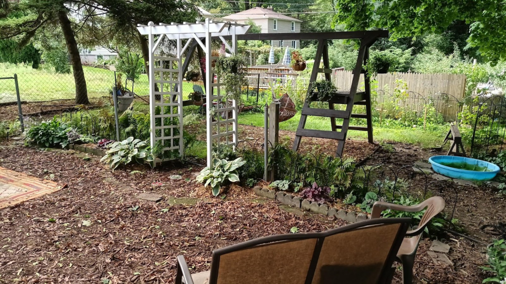 Backyard view showing a white garden arch, planted beds, swingset repurposed with hanging baskets, and a small blue plastic wading pool.